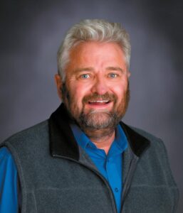 A middle-aged man with gray hair and a beard smiles at the camera. He is wearing a blue shirt and a gray fleece vest, posing in front of a neutral, dark gray background.