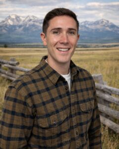 A young man in a brown plaid shirt smiles while standing in front of a wooden fence on one of the scenic Montana ranches, with snow-capped mountains and a partly cloudy sky in the background.