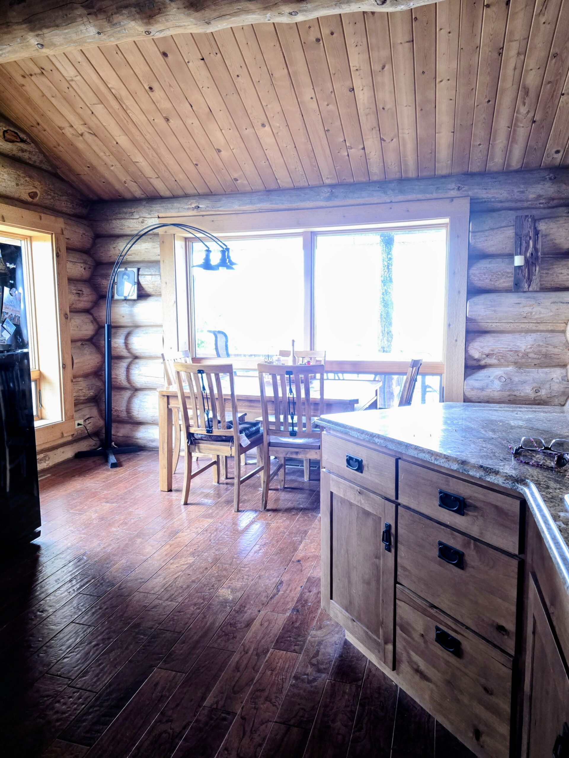 A sunlit dining area in a log cabin features a wooden table with four chairs near large windows, rustic wood walls, and hardwood floors—capturing the inviting warmth often found in Montana ranches or farms for sale. Part of the kitchen counter is visible in the foreground.