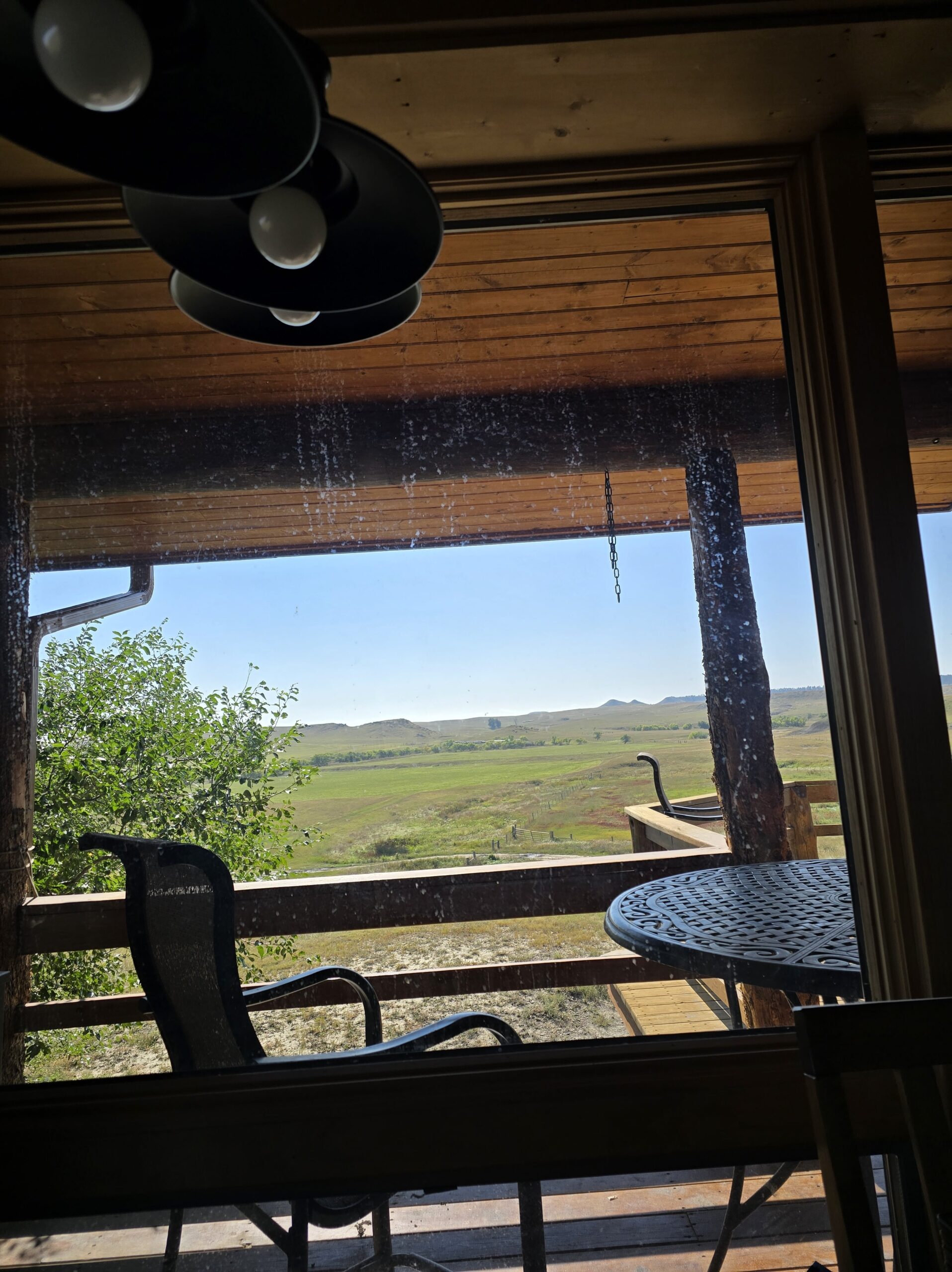 View through a window showing a covered wooden porch with a round metal table, chairs, and scenic green fields and hills—perfect for those seeking farms for sale or hunting land. A hanging lamp is partly visible inside.