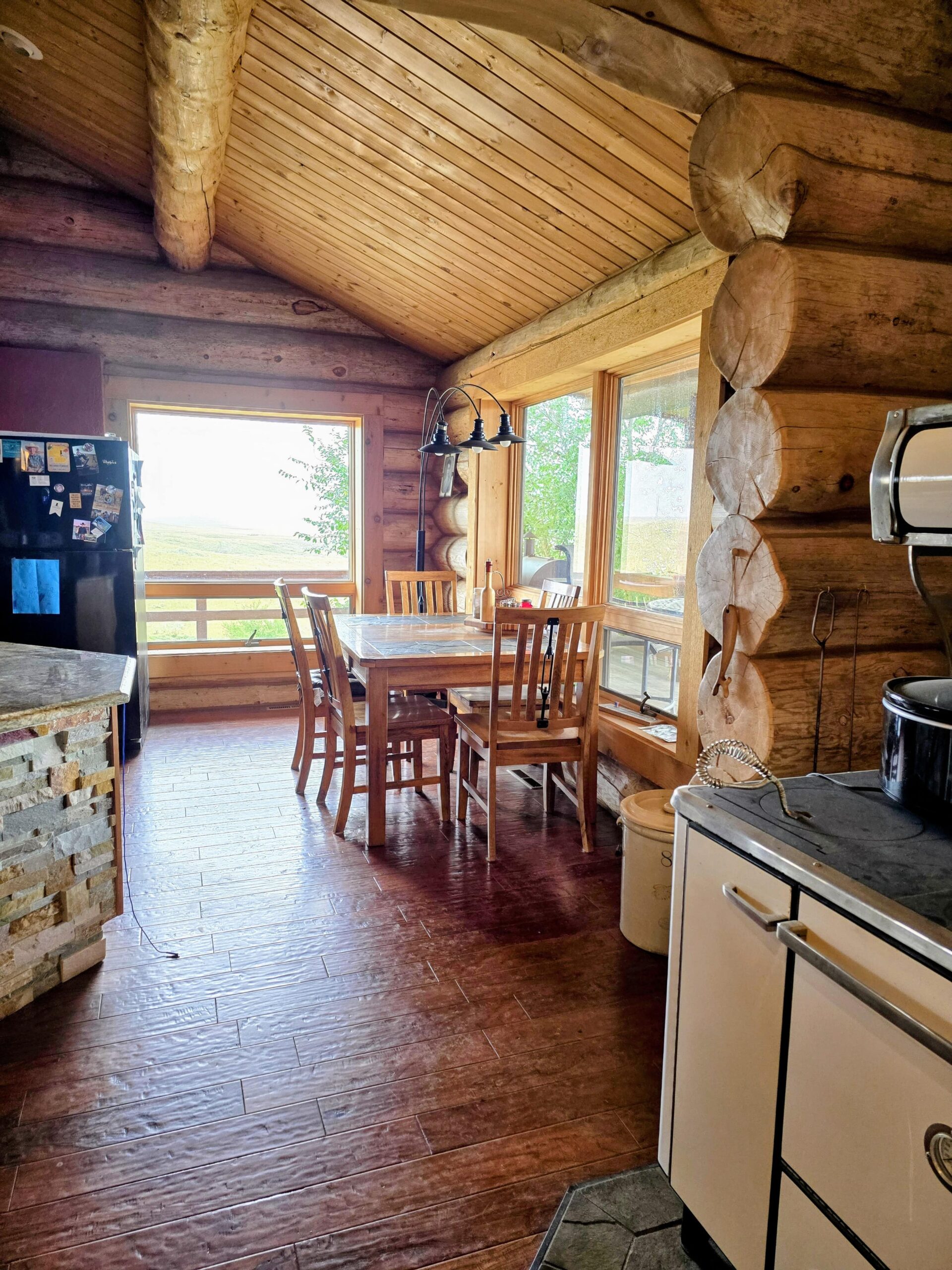 A cozy log cabin kitchen with wooden walls, a dining table and chairs by large windows, a black refrigerator, and a vintage white stove. Natural light fills the space, highlighting rustic decor often found in classic Montana ranches.