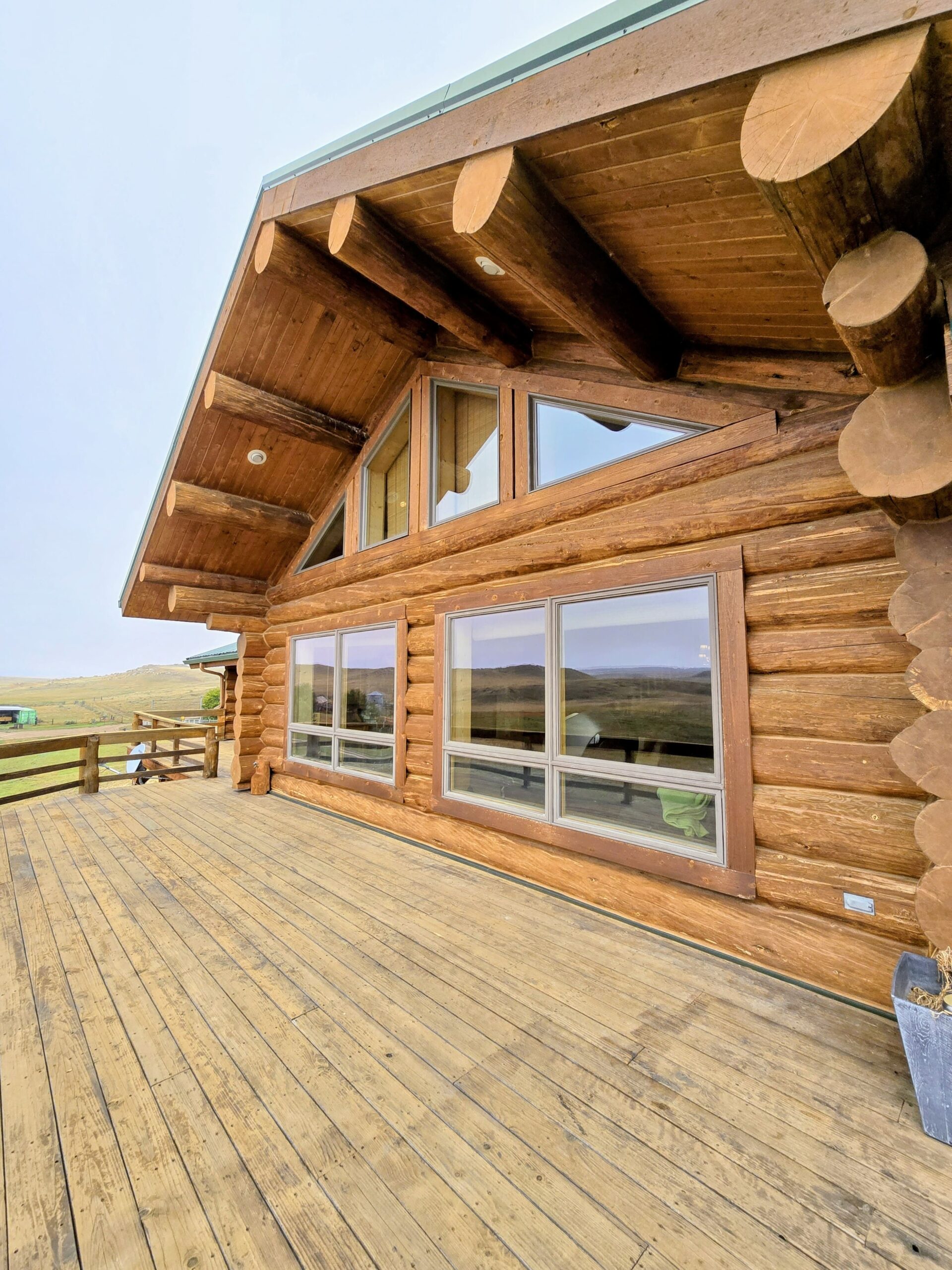 A wooden deck fronts a log cabin with large windows and a triangular roof, set in an open, grassy landscape—perfect for those seeking cozy ranch homes under a light, overcast sky.