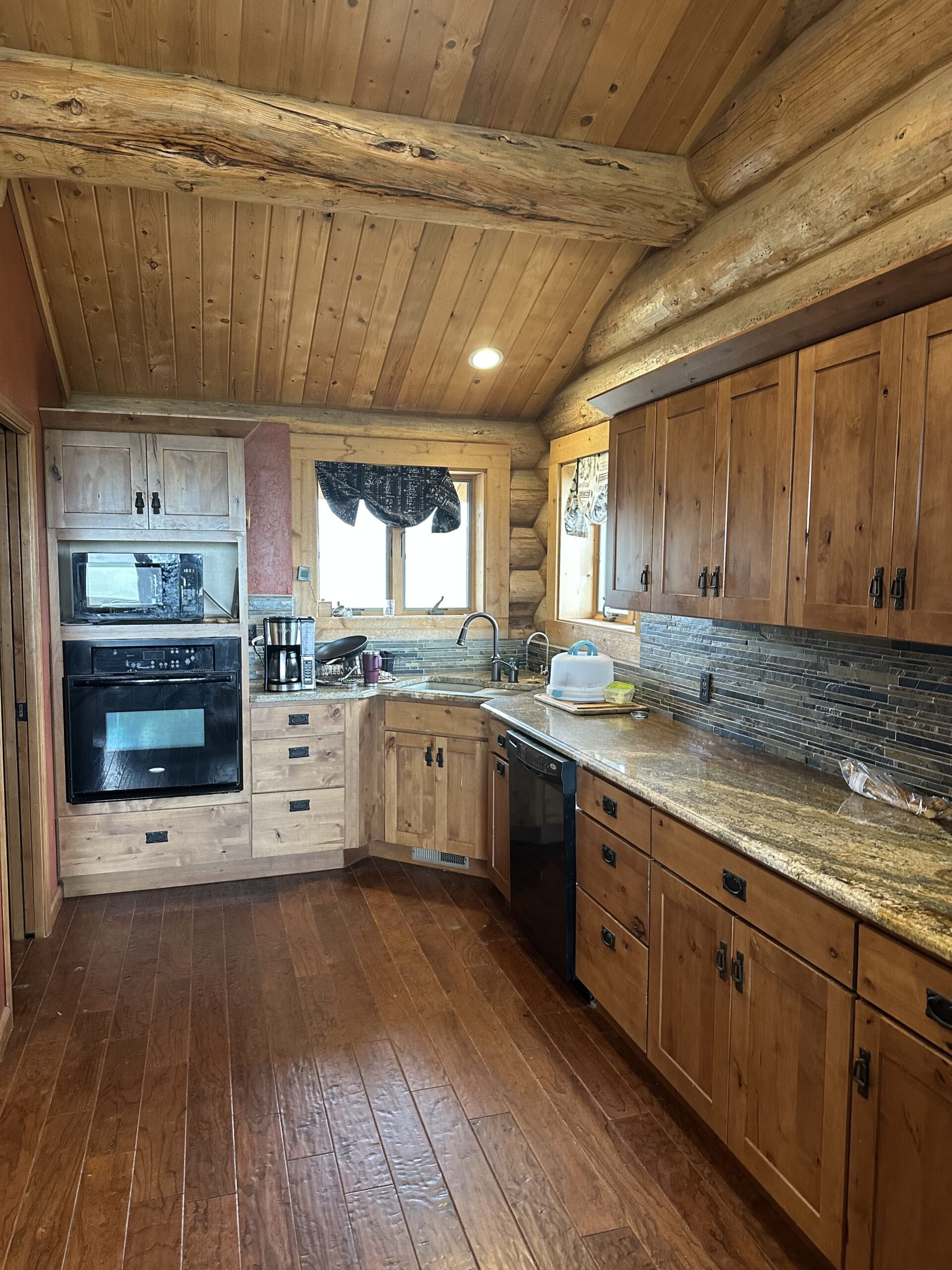 A rustic log cabin kitchen with wood cabinets, granite countertops, stainless steel appliances, a double oven, tile backsplash, and wood flooring—perfect for ranch homes or those seeking the charm of country living. Light streams in through a window above the sink.