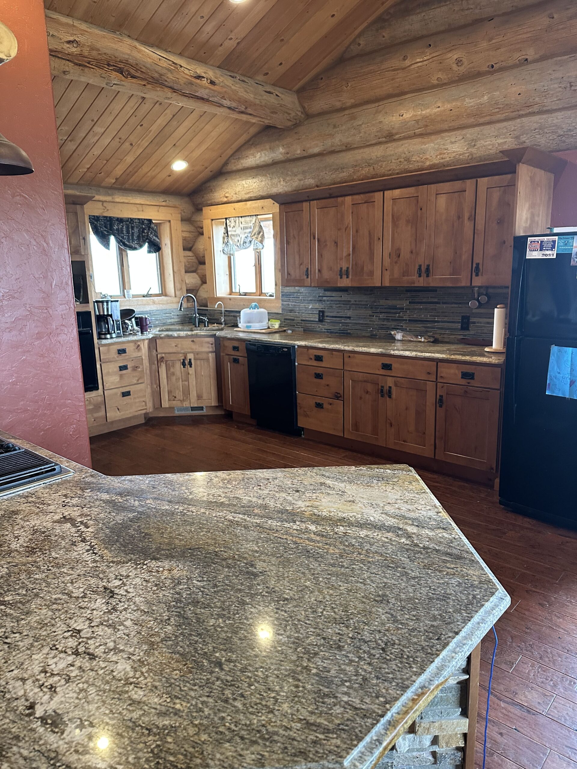 A rustic kitchen with wooden cabinets, log walls, a stone countertop, black appliances, a window with curtains, and a sloped wooden ceiling with exposed beams—perfect for ranch homes or cozy Montana ranches.
