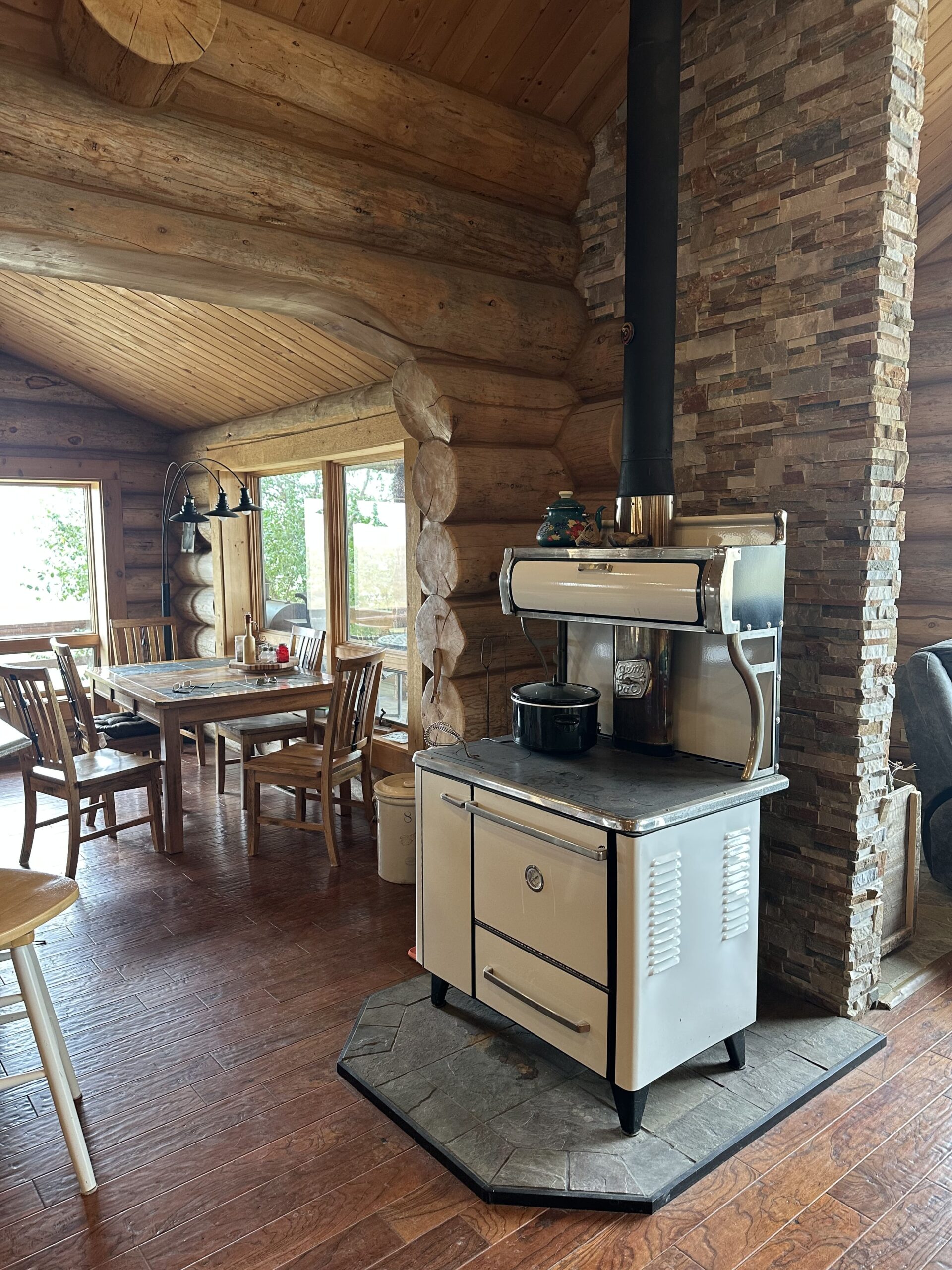 A vintage white wood-burning stove sits on a stone platform in a cozy log cabin kitchen, perfect for classic ranch homes, with wooden floors, a dining table, and large windows letting in natural light.