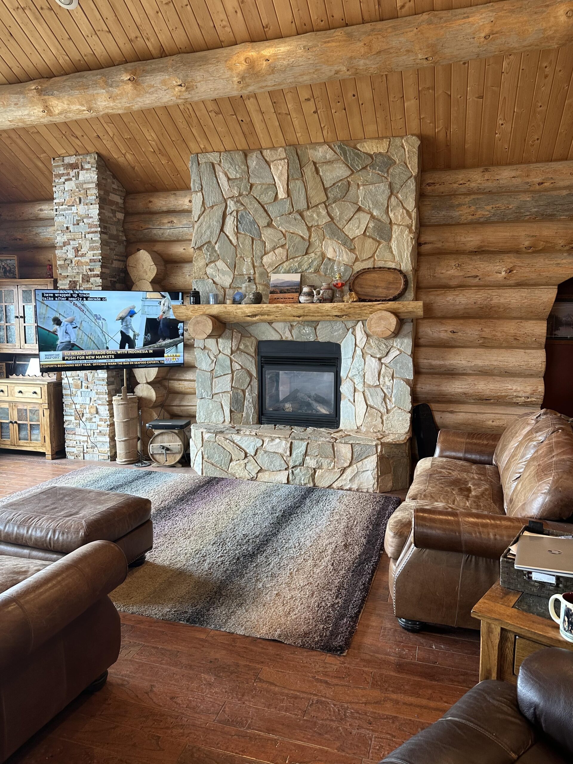 A cozy log cabin living room with a stone fireplace, leather sofas, wooden floors, and a large rug creates a warm, rustic retreat—perfect inspiration for those searching for ranches for sale or dreaming of country comfort.