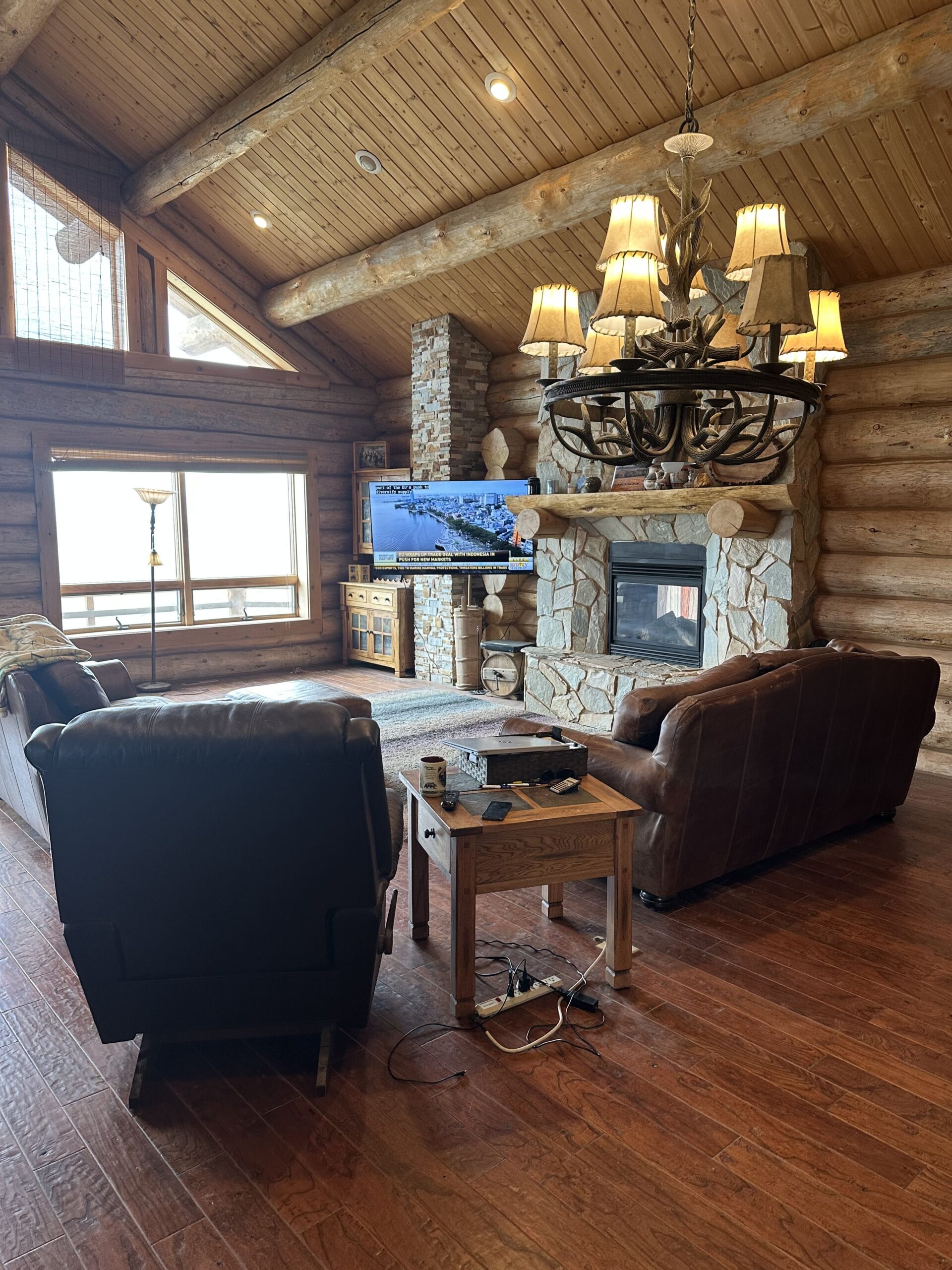 A cozy log cabin living room with leather sofas, a stone fireplace, a large antler chandelier, and a TV above the hearth. Wooden floors and walls create a rustic, warm atmosphere reminiscent of Montana ranches.