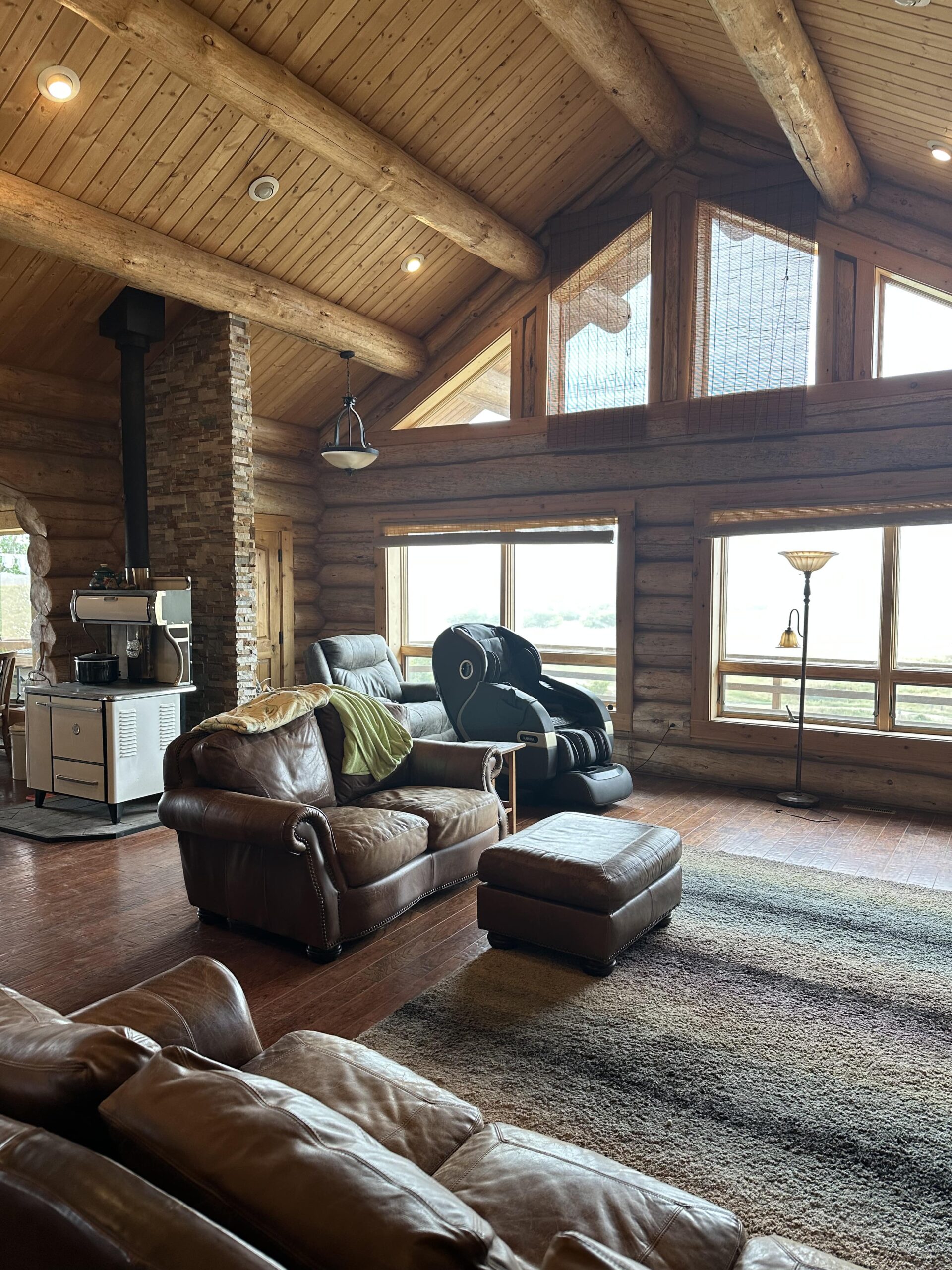 A cozy log cabin living room with leather chairs, a matching ottoman, a rustic stove, large windows letting in natural light, and wooden beam ceilings—perfect for relaxing after exploring nearby Montana ranches or hunting land.