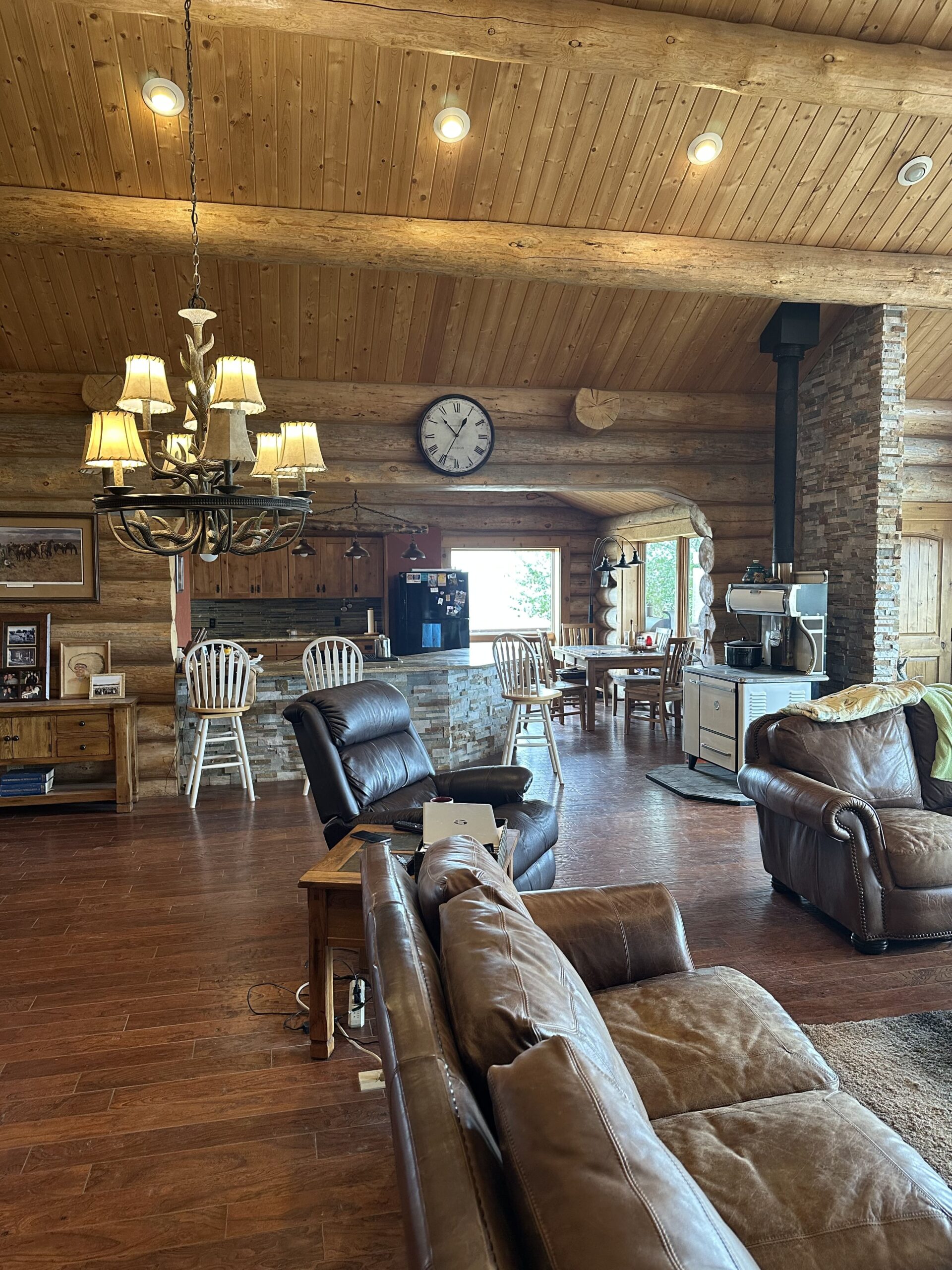 A cozy log cabin living area with leather sofas, a rustic chandelier, a kitchen island with white chairs, and wood-paneled walls. This inviting space is perfect for those seeking ranch homes or browsing ranches for sale. Natural light fills the room.