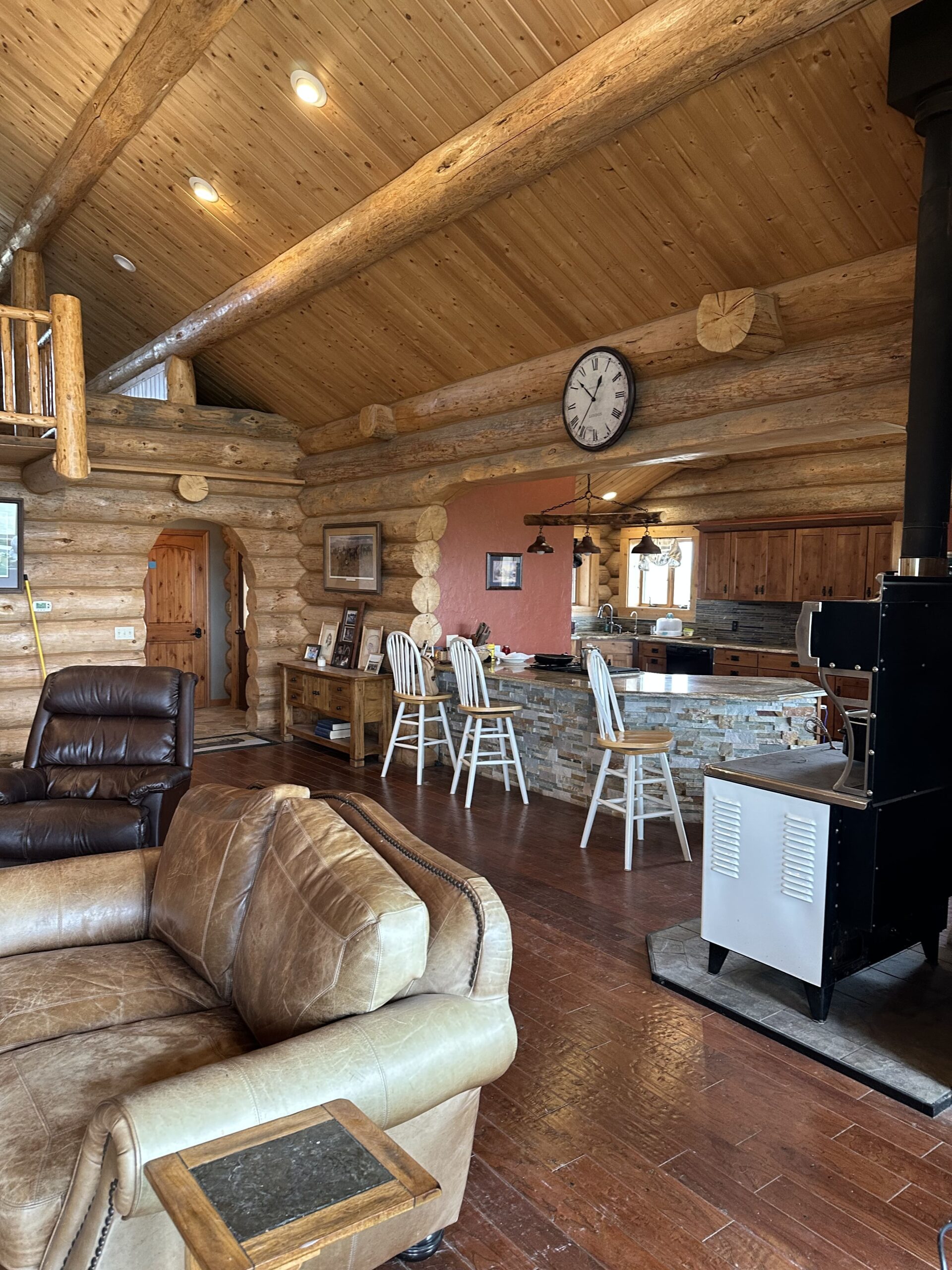 A cozy log cabin interior with wood beams, leather furniture, a stone kitchen island with barstools, a large clock on the wall, and a wood-burning stove on a tile hearth—perfect for those seeking ranch homes.