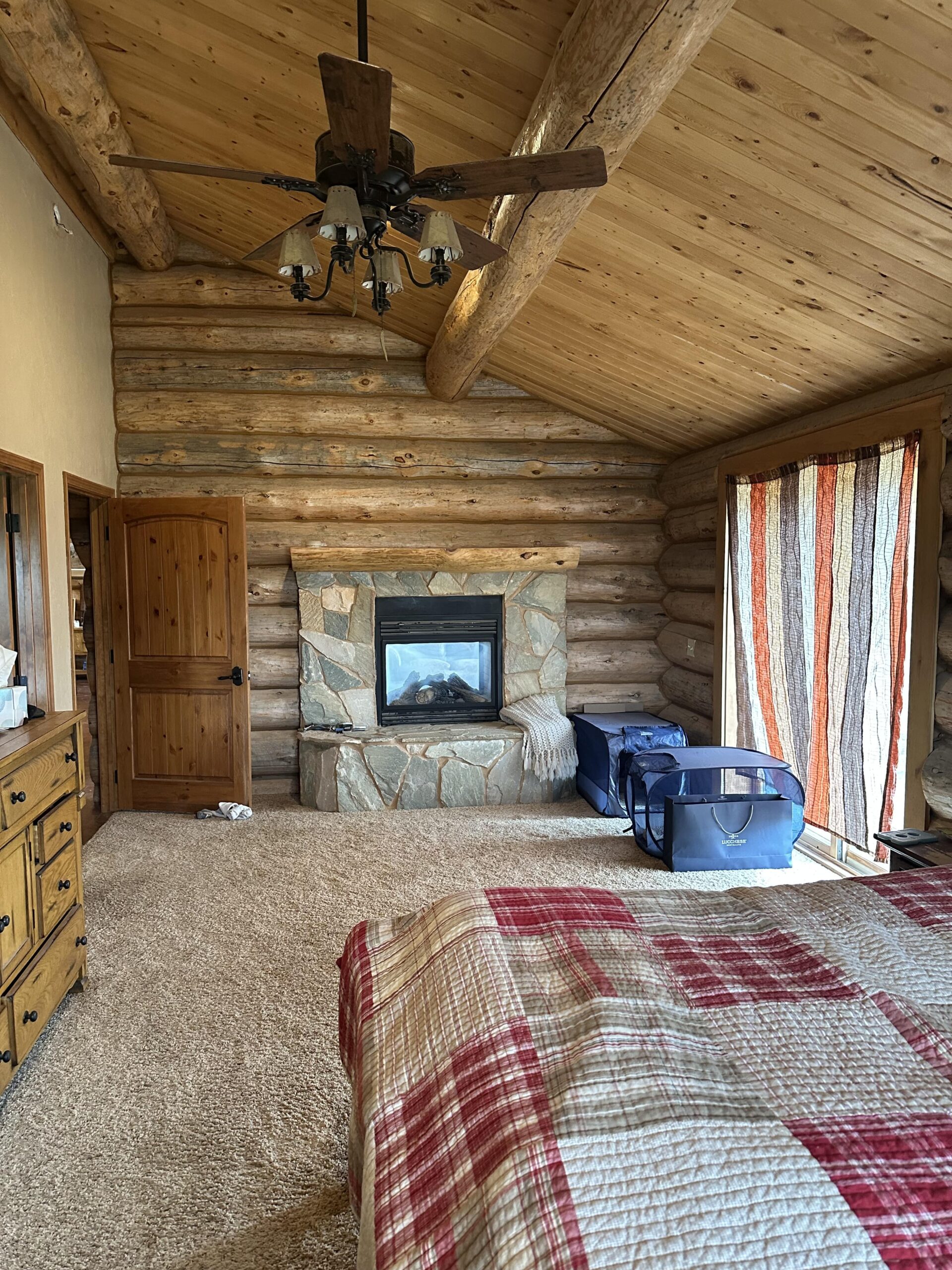 A cozy log cabin bedroom reminiscent of classic ranch homes, with a stone fireplace, wood-paneled ceiling, a patchwork quilt on the bed, wooden furniture, and striped curtains letting in sunlight. A suitcase sits by the window.