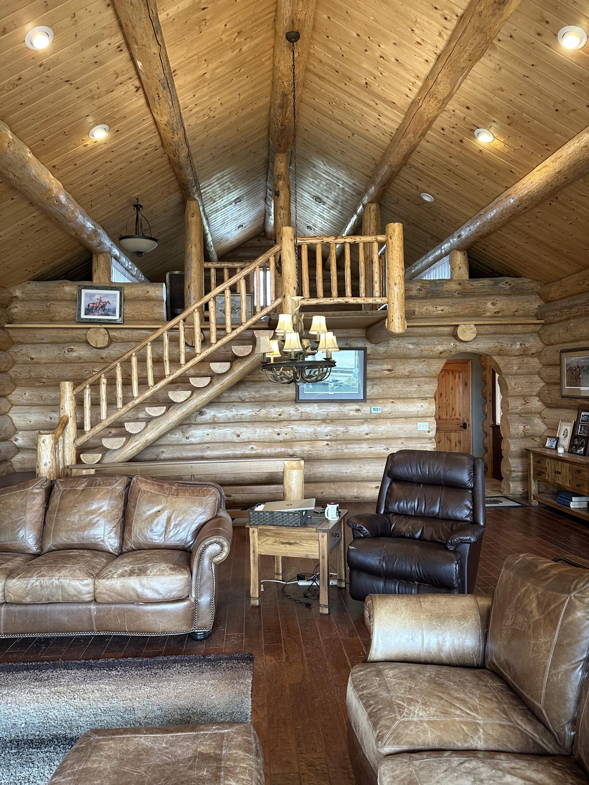 A cozy log cabin living room with wood walls and ceiling, leather sofas and recliners, a staircase leading to a loft, a chandelier, and framed photos—perfect inspiration for those seeking Montana ranches or rustic hunting land retreats.