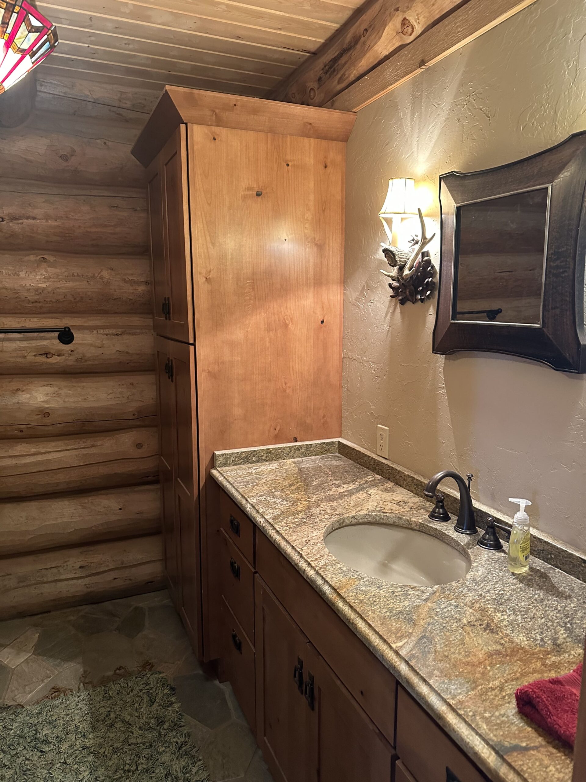 A rustic bathroom with log walls, a wooden cabinet, a granite countertop with a sink, an ornate mirror, a sconce light, and a red towel evokes the timeless charm found in classic ranch homes.