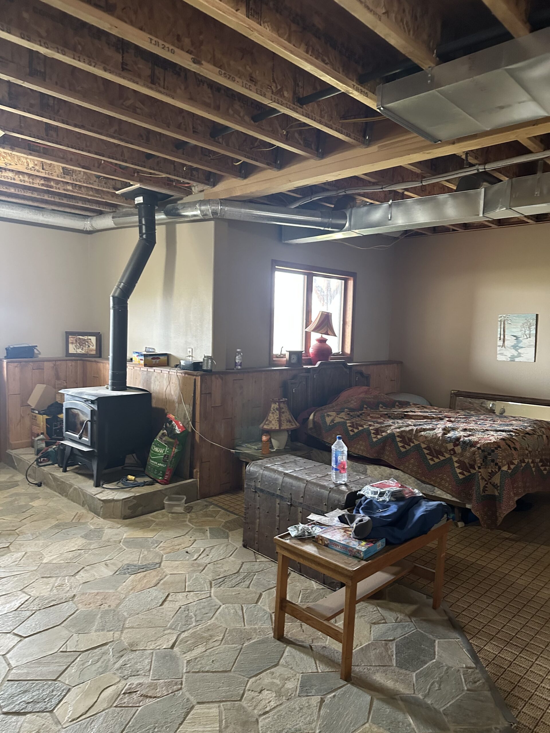 A cozy basement room reminiscent of Montana ranches, with exposed wooden beams, a wood-burning stove, a patchwork quilt on the bed, a wooden chest, and a small table on stone-tiled floors. Sunlight filters through the window.