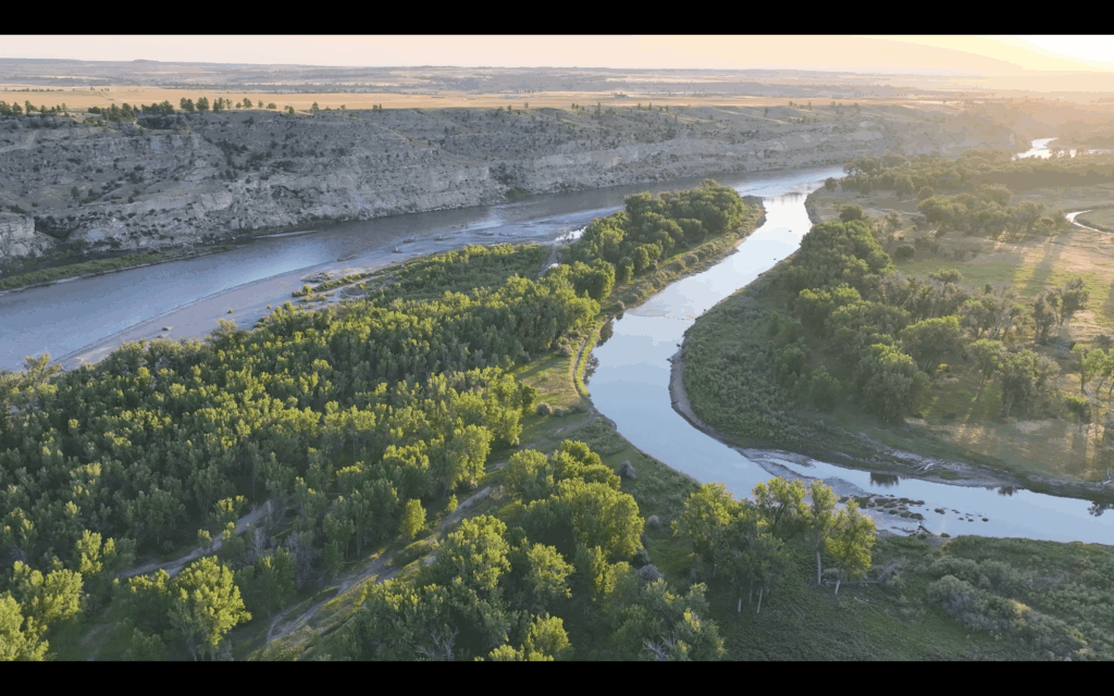 A river winds through a green landscape with trees and shrubs, bordered by rocky cliffs and open plains—idyllic scenery often found near ranches for sale—under a soft, golden sunset sky.