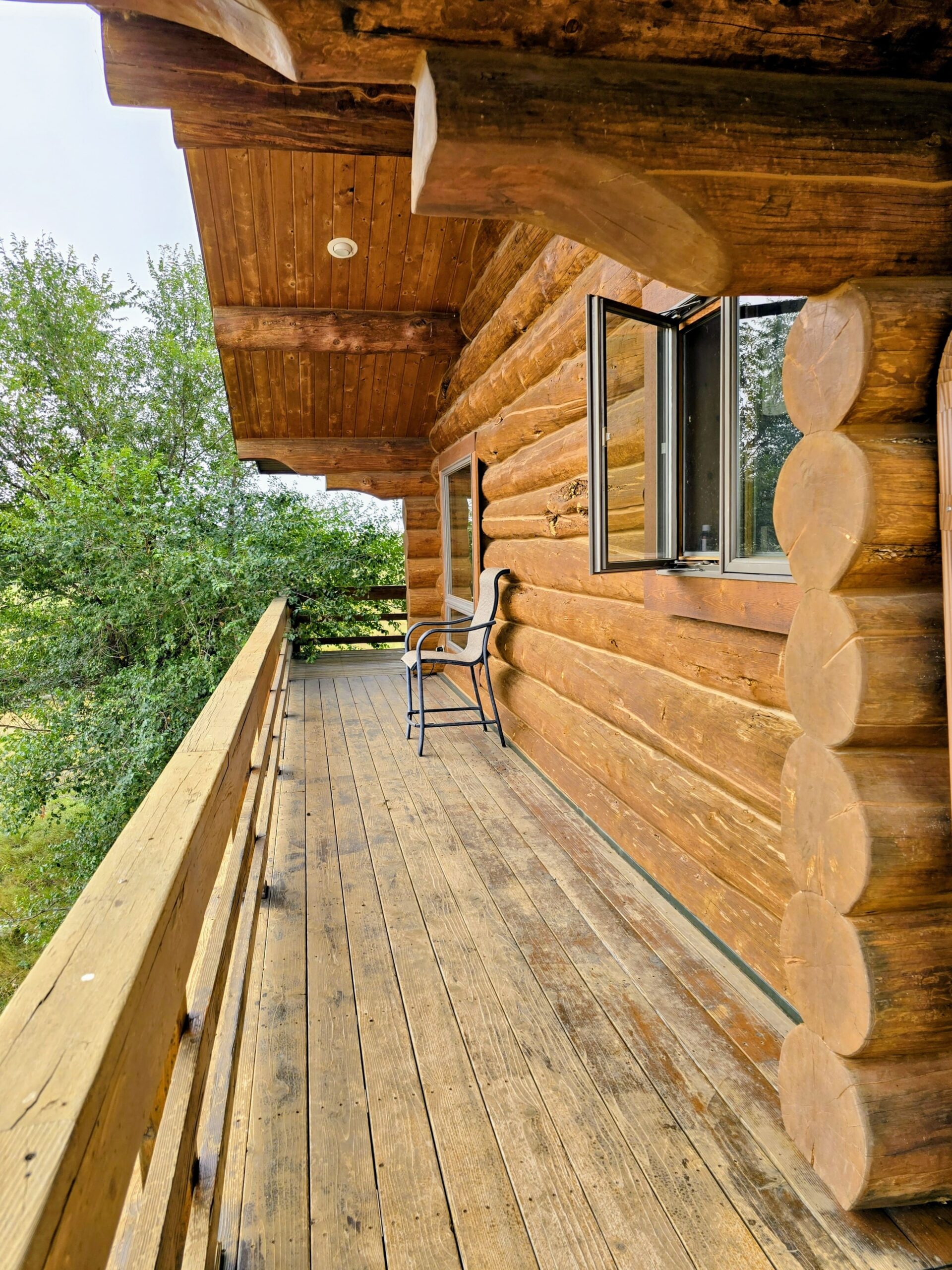 A wooden balcony with a rustic log cabin wall, an open window, and a single chair overlooks green trees on a cloudy day—capturing the peaceful charm often found on Montana ranches.