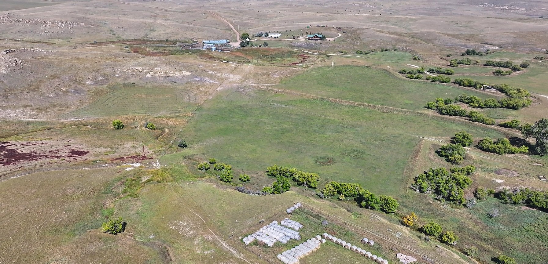 Aerial view of a vast, open landscape with grassy fields, patches of trees, and scattered buildings—ideal for hunting land or ranch homes. A cluster of small white tents sits in the foreground, with rolling hills visible in the distance.