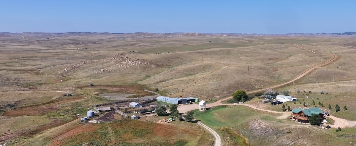 Aerial view of a rural ranch with several buildings, fenced areas, and a winding dirt road surrounded by vast, rolling grasslands under a clear blue sky—perfect for those seeking Montana ranches or hunting land.
