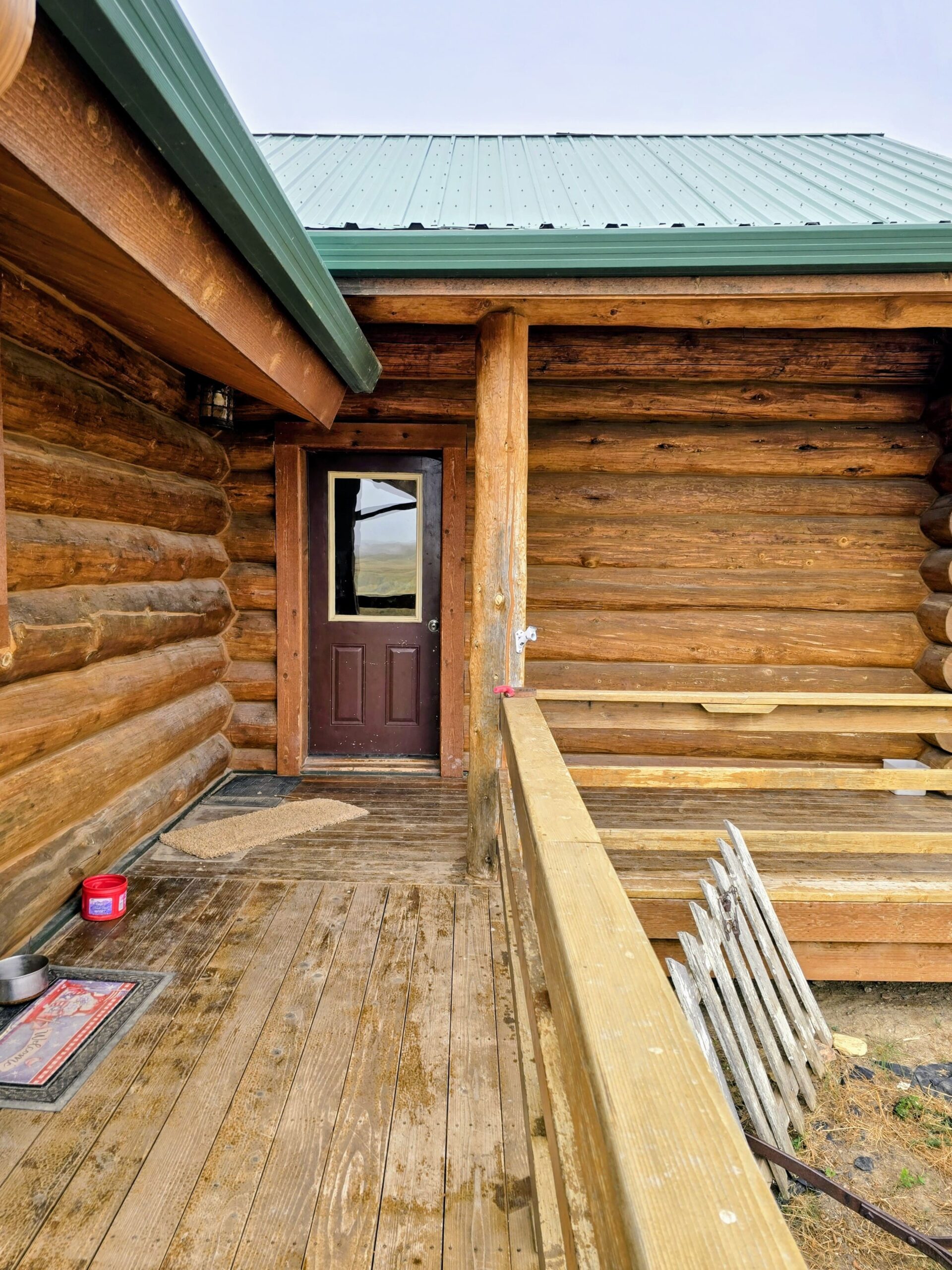 A wooden deck leads to a brown door on a log cabin with a green metal roof, common in classic ranch homes. A bench, pet bowl, and red container sit on the weathered deck, revealing signs of use.