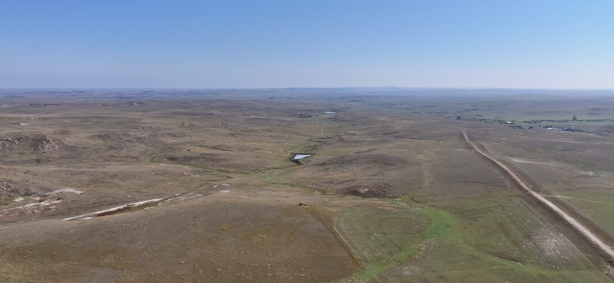 Aerial view of a vast, dry landscape with rolling hills, sparse vegetation, a small pond near the center, and a dirt road stretching into the distance—classic scenery often found among Montana ranches under a clear blue sky.