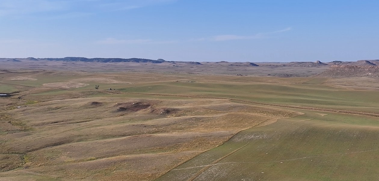 A vast, open grassland under a clear blue sky, with gently rolling hills and distant rocky outcroppings on the horizon—classic scenery found with Montana ranches and prime hunting land, expansive and dry with minimal vegetation.