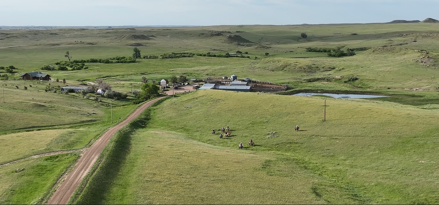 A rural landscape with a dirt road leading to a farm surrounded by green fields and rolling hills. Several people on horseback ride along a grassy hill, showcasing the beauty of Montana ranches for sale. Scattered buildings and a pond are visible in the distance.