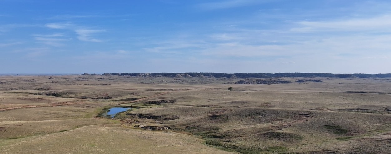 A vast, open prairie landscape under a blue sky, with rolling hills, a small pond, and sparse vegetation scattered across the dry grasslands—perfect for those seeking hunting land or dreaming of ranch homes.