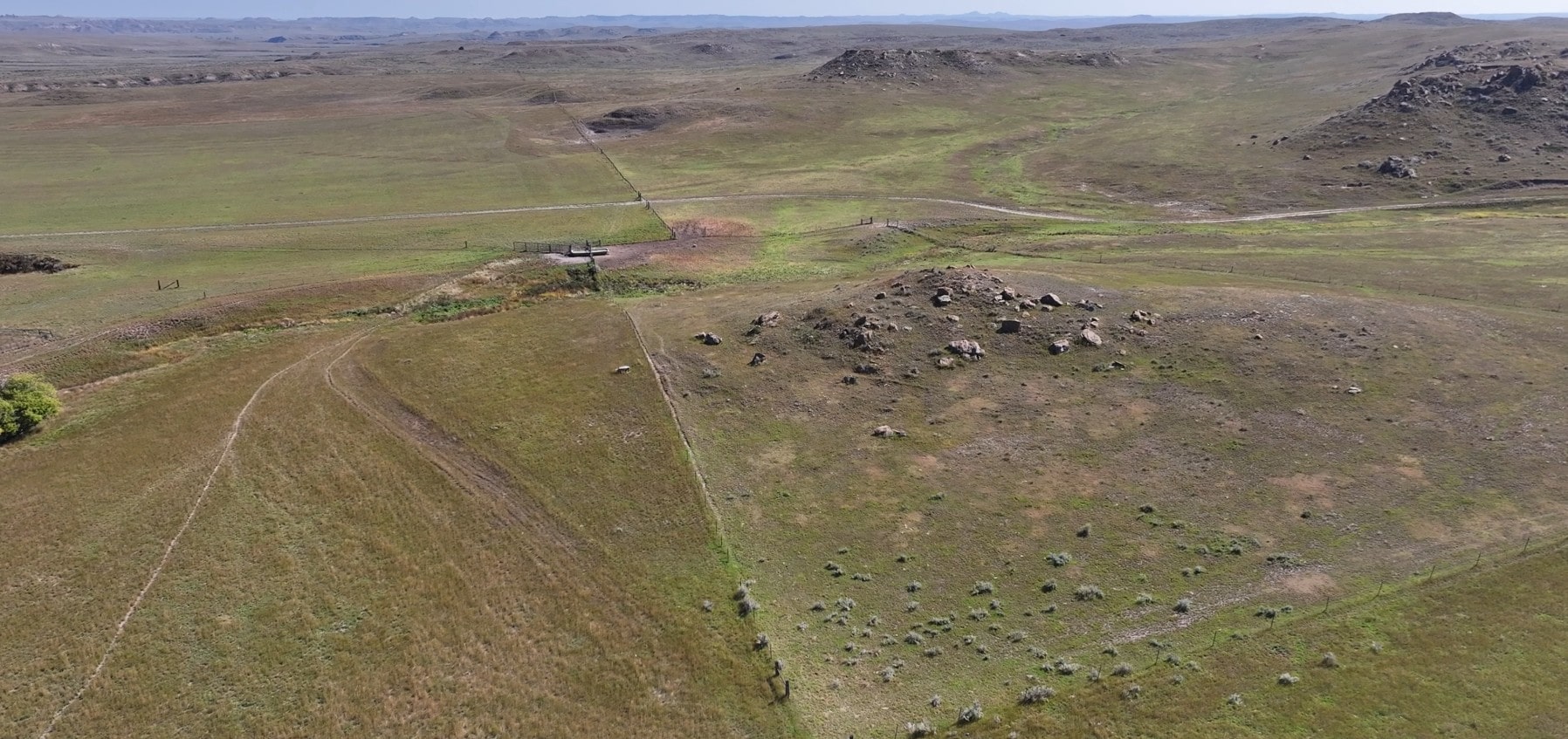 Aerial view of a vast grassy plain with low rolling hills, scattered rocks, and dirt roads—ideal for hunting land; a small structure sits near the center left, showcasing the potential of Montana ranches.