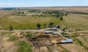 Aerial view of a rural ranch with fenced enclosures, small buildings, and scattered trees, surrounded by vast open fields under a blue sky—ideal for those seeking ranches for sale.