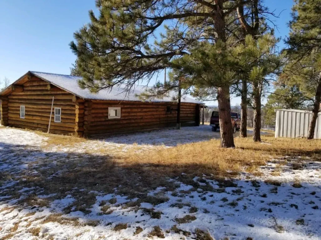 A rustic log cabin sits among pine trees on a patchy, snow-covered yard under a clear blue sky—an inviting scene reminiscent of classic montana ranches. A metal shed and parked vehicle are visible near the cabin.