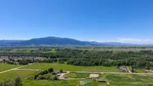 A wide landscape view of green fields, scattered trees, and a few ranch homes in the foreground, with distant mountains under a clear blue sky.