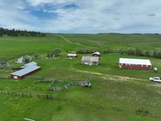 Aerial view of a rural farm with several buildings, fenced areas, a white truck, and green fields under a partly cloudy sky—ideal for those seeking Montana ranches. Rolling hills are visible in the background.