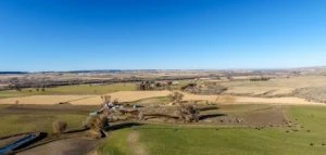 A wide view of open farmland under a clear blue sky, with fields, scattered trees, and a few ranches for sale visible in the distance.