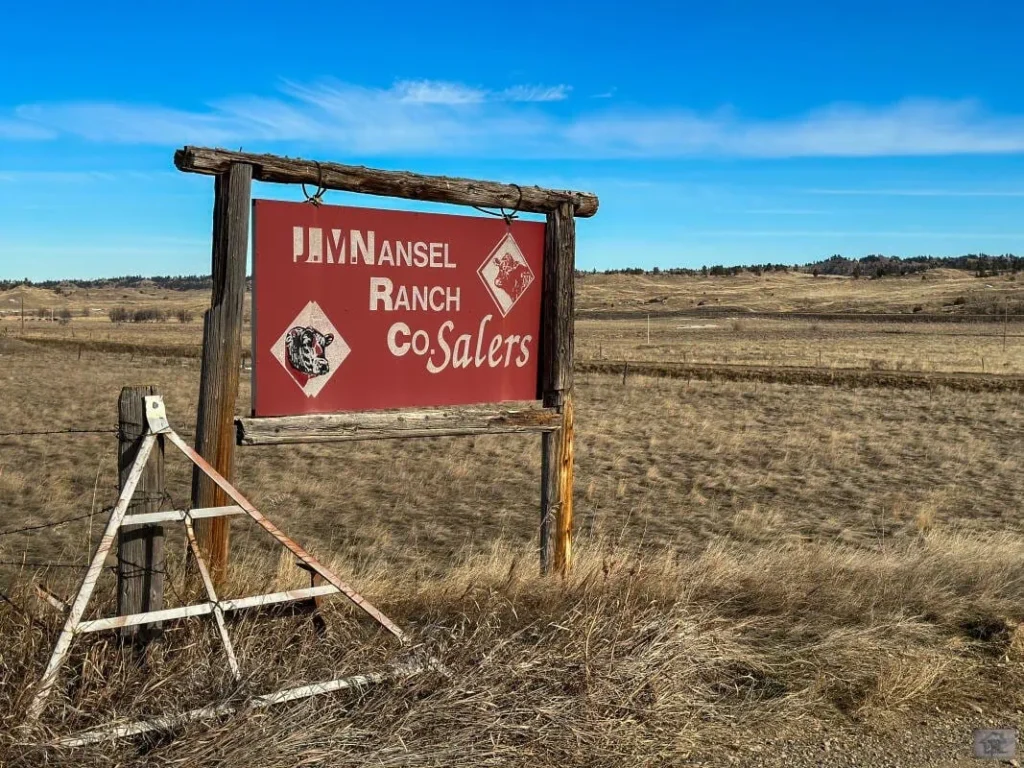 A weathered red sign reading Mansel Ranch Co. Salers with images of cattle stands in a dry, grassy field under a blue sky—an inviting scene for those seeking ranches for sale amid rolling hills and sparse trees.