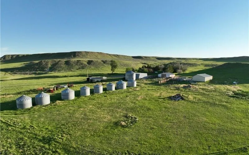 Aerial view of a rural farm with multiple silver grain silos, barns, equipment, and green grassy fields—one of the picturesque Montana ranches surrounded by rolling hills under a clear blue sky.