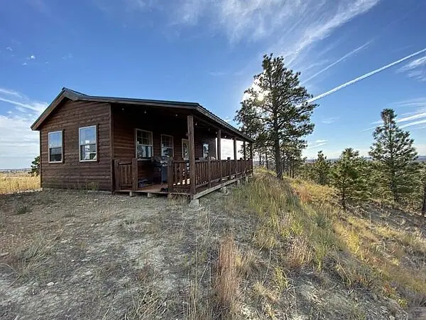 A small wooden cabin with a covered porch sits on a grassy hilltop, reminiscent of classic ranch homes, surrounded by pine trees under a blue sky with wispy clouds and sun rays.