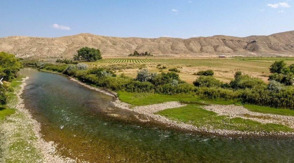 A winding river flows through green fields and rows of crops, with scattered trees and ranch homes, set against dry, rolling hills under a clear blue sky.