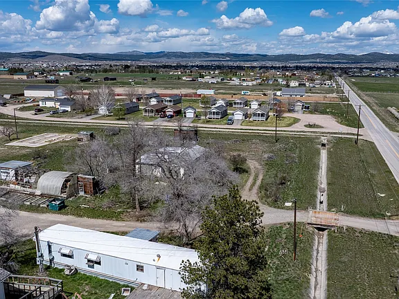Aerial view of a rural area with scattered homes, open fields, dirt roads, a highway, and distant hills under a partly cloudy blue sky.