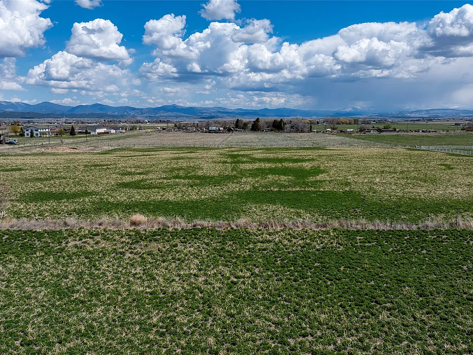 A wide, open grassy field under a partly cloudy sky with distant mountains and a few scattered buildings visible along the horizon.