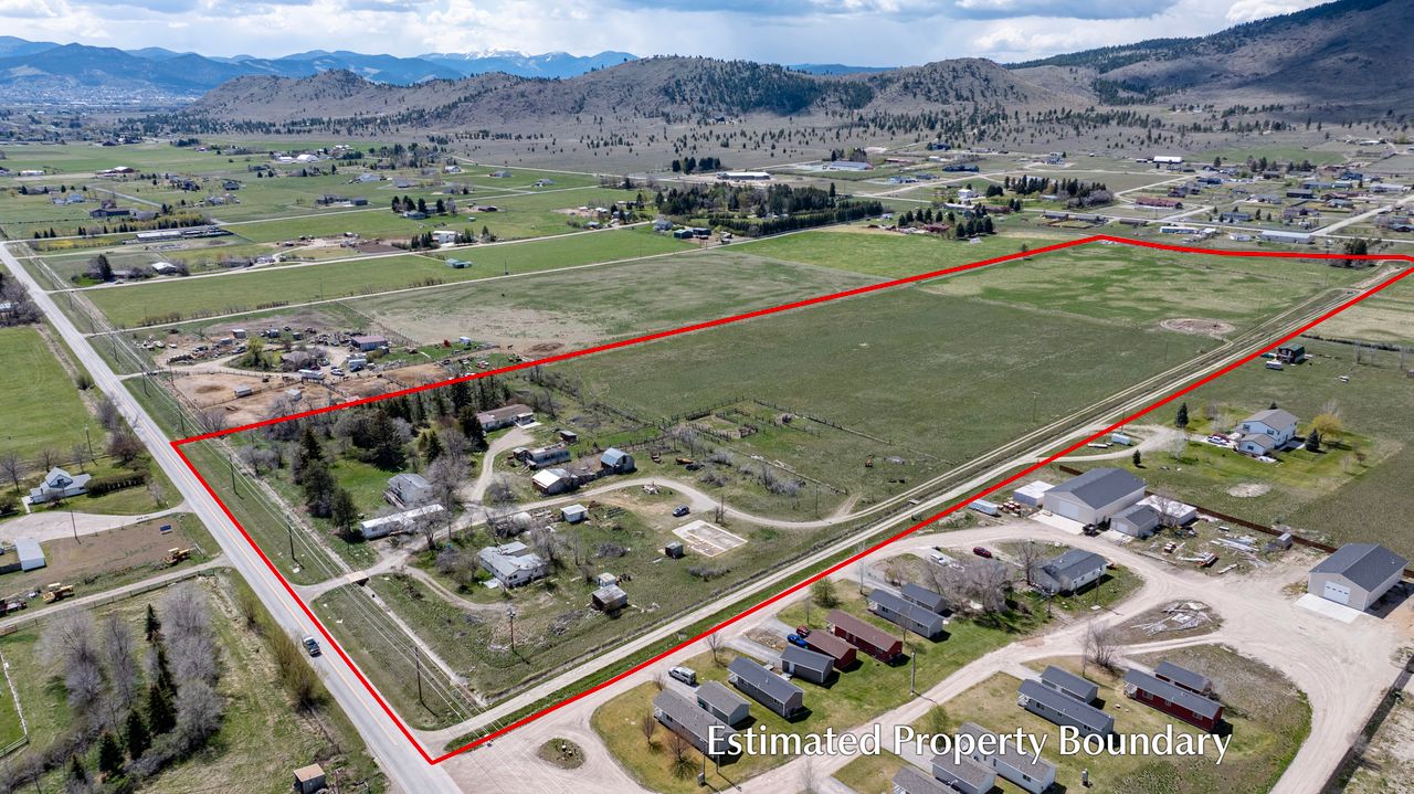 Aerial view of a large, mostly grassy property outlined in red, showing farmhouses, roads, and nearby homes with mountains in the background. Text reads Estimated Property Boundary.