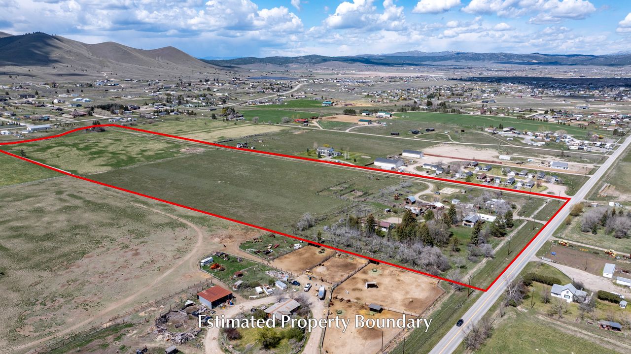Aerial view of a large rural property outlined in red, showing fields, houses, and nearby roads under a partly cloudy sky with distant hills in the background. Text at bottom reads, Estimated Property Boundary.