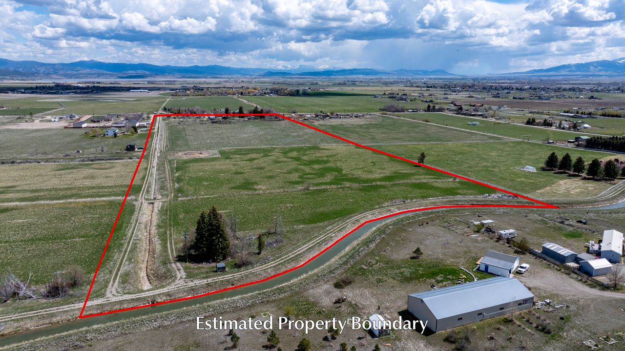 Aerial view of a large green field bordered with a red line showing the estimated property boundary, with scattered buildings, trees, and distant mountains under a cloudy sky.