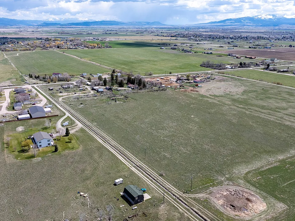 Aerial view of a rural landscape with scattered houses, barns, green fields, dirt roads, and distant mountains beneath a cloudy sky.