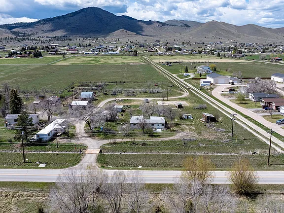 Aerial view of a rural area with scattered houses, green fields, and a road in the foreground. Rolling hills and mountains are visible in the background under a partly cloudy sky.