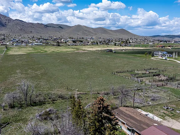 A wide view of a rural landscape with open grassy fields, a few scattered houses, fenced areas, and rolling hills in the background under a partly cloudy sky.