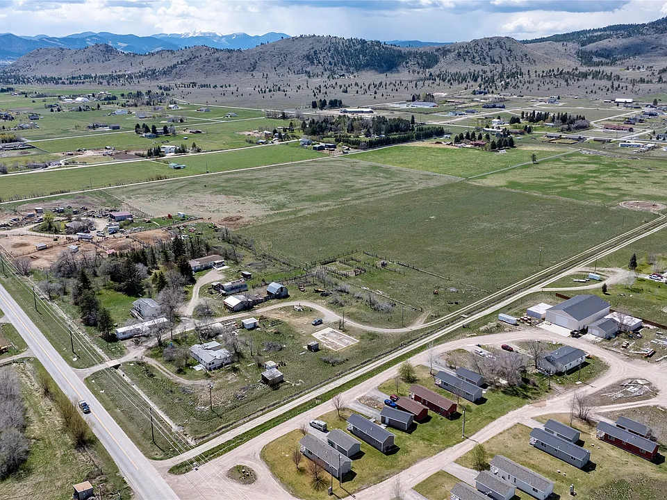 Aerial view of a rural area with scattered houses, barns, open green fields, winding roads, and distant hills under a partly cloudy sky.