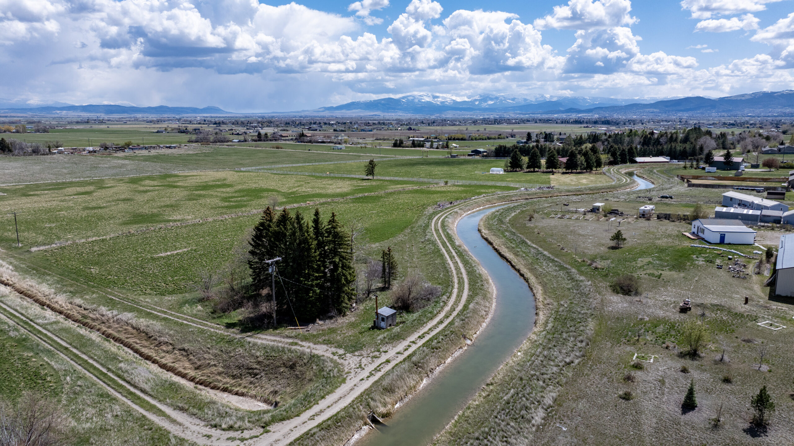 A winding irrigation canal cuts through green farmland under a partly cloudy sky, with scattered buildings, trees, and distant snow-capped mountains in the background.