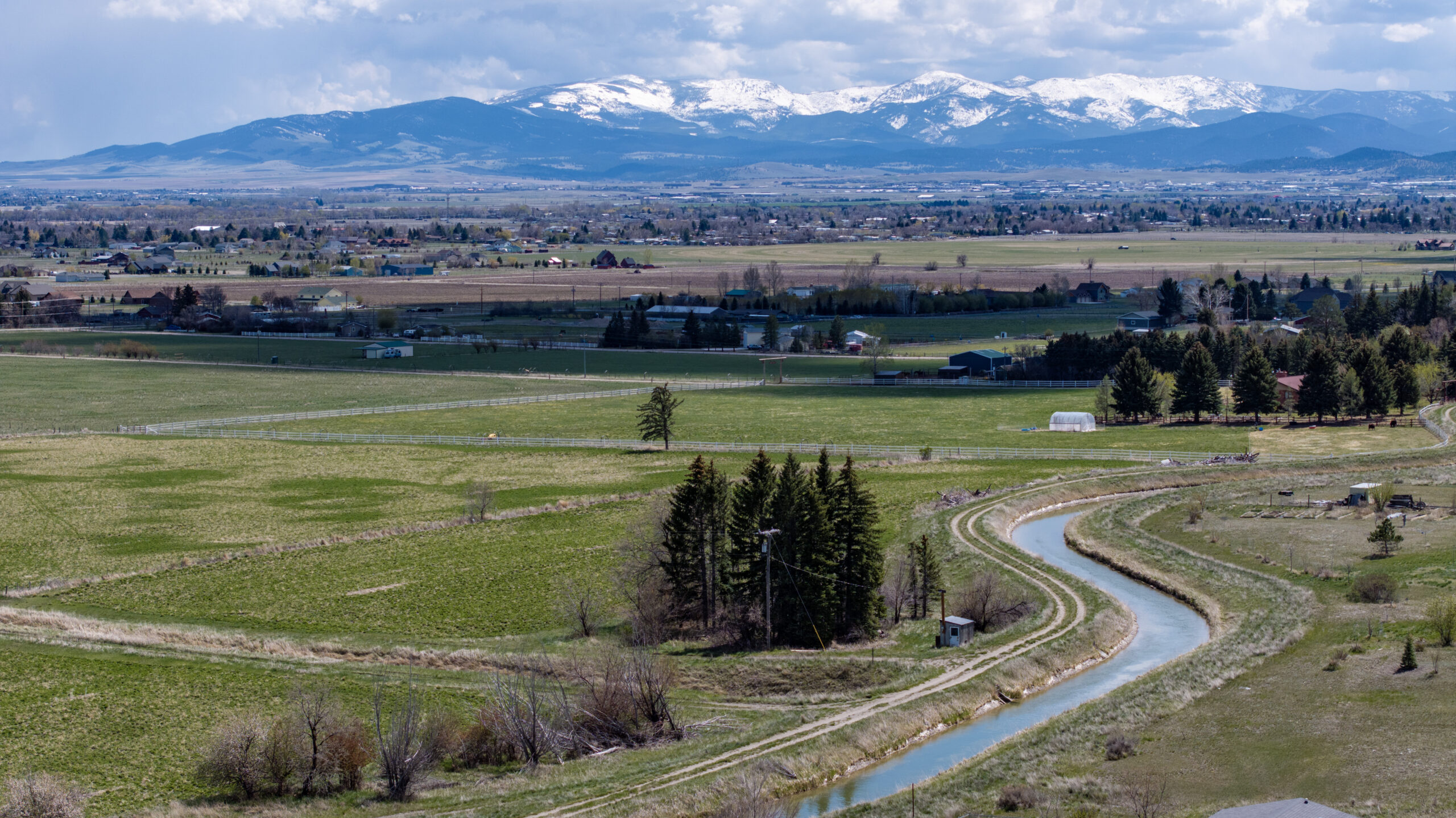 A winding river flows through green fields and farmland with scattered houses and trees; snow-capped mountains rise in the distance under a partly cloudy sky.