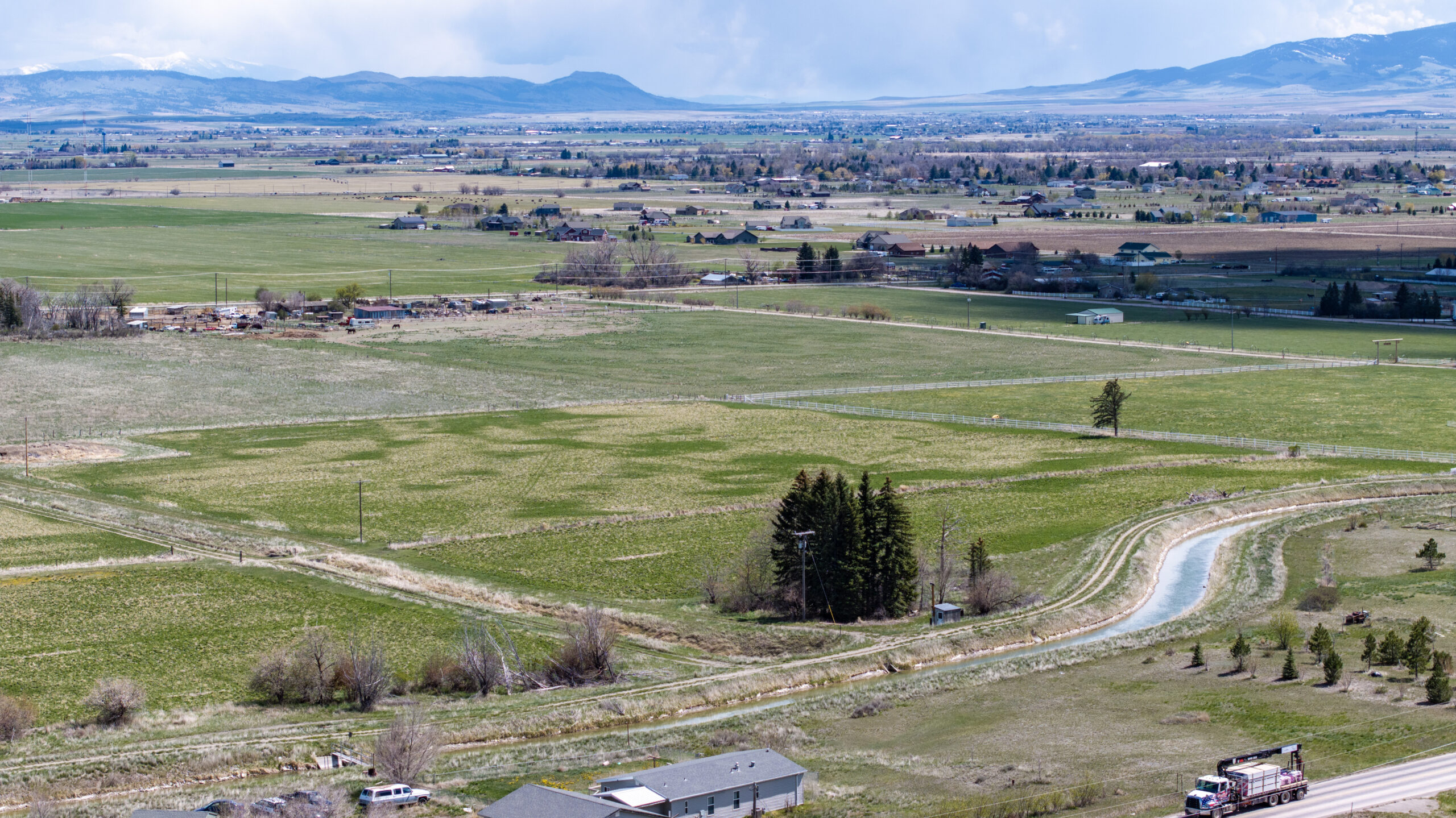 A wide view of green farmland with scattered houses, trees, and a canal winding through the fields, set against distant mountains under a partly cloudy sky.