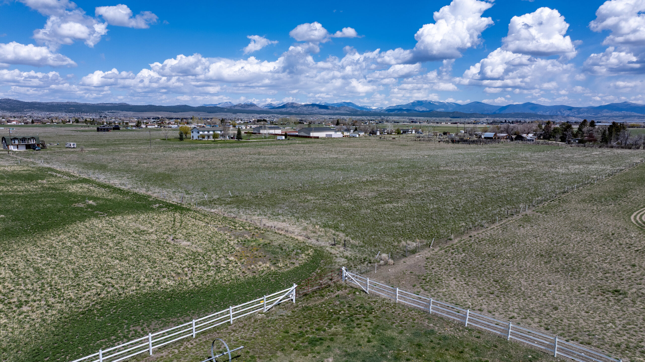 Aerial view of a wide, open rural field with patchy green and brown grass, bordered by white fences, scattered houses, and distant mountains under a blue sky with fluffy clouds.